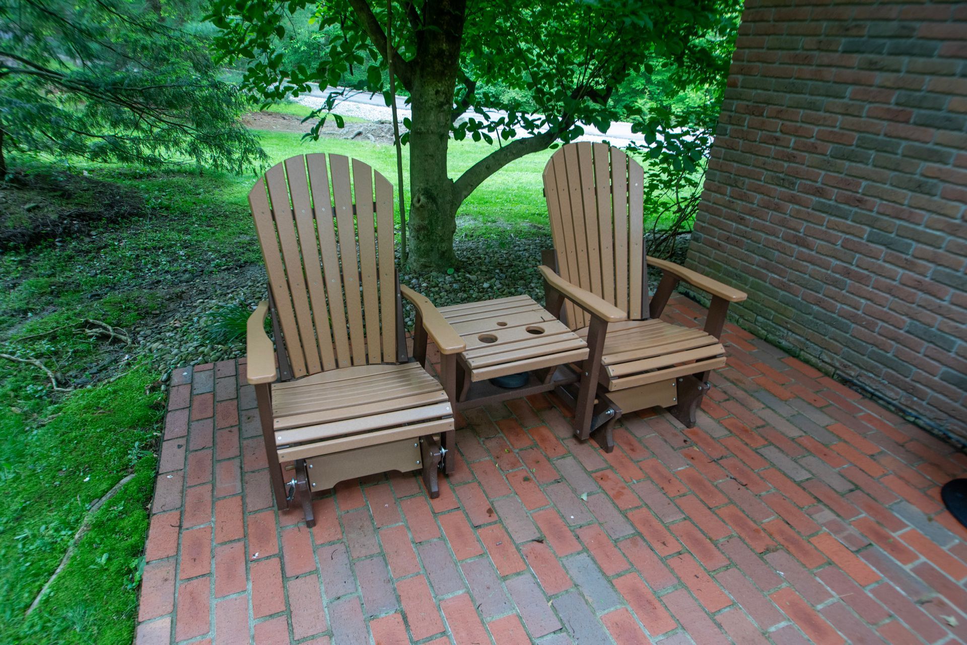 Two wooden chairs and a small table on a brick patio, next to a brick wall and greenery.