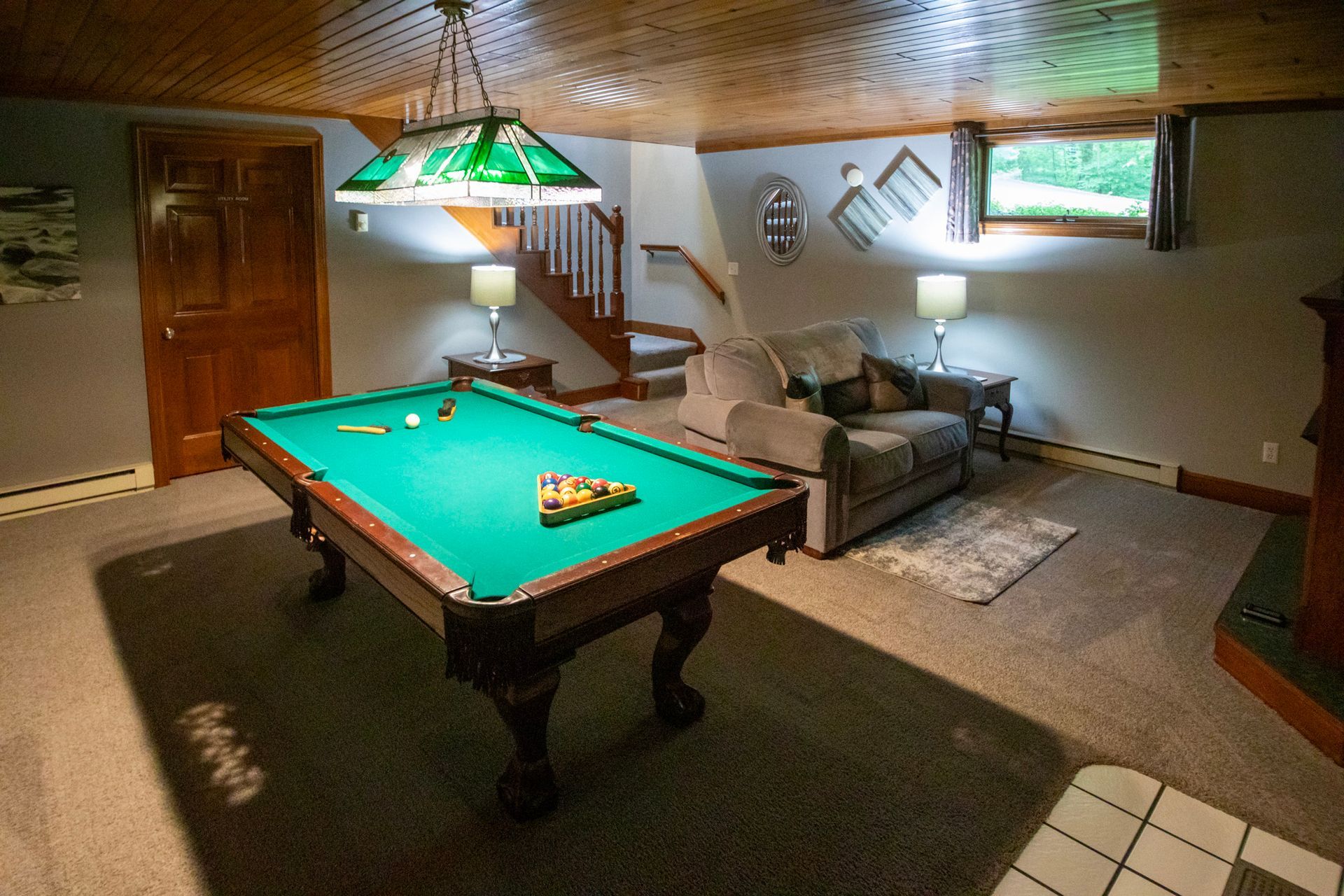 Pool table in a finished basement, lit by a green-shaded lamp. Couch, stairs, and small windows are also present.