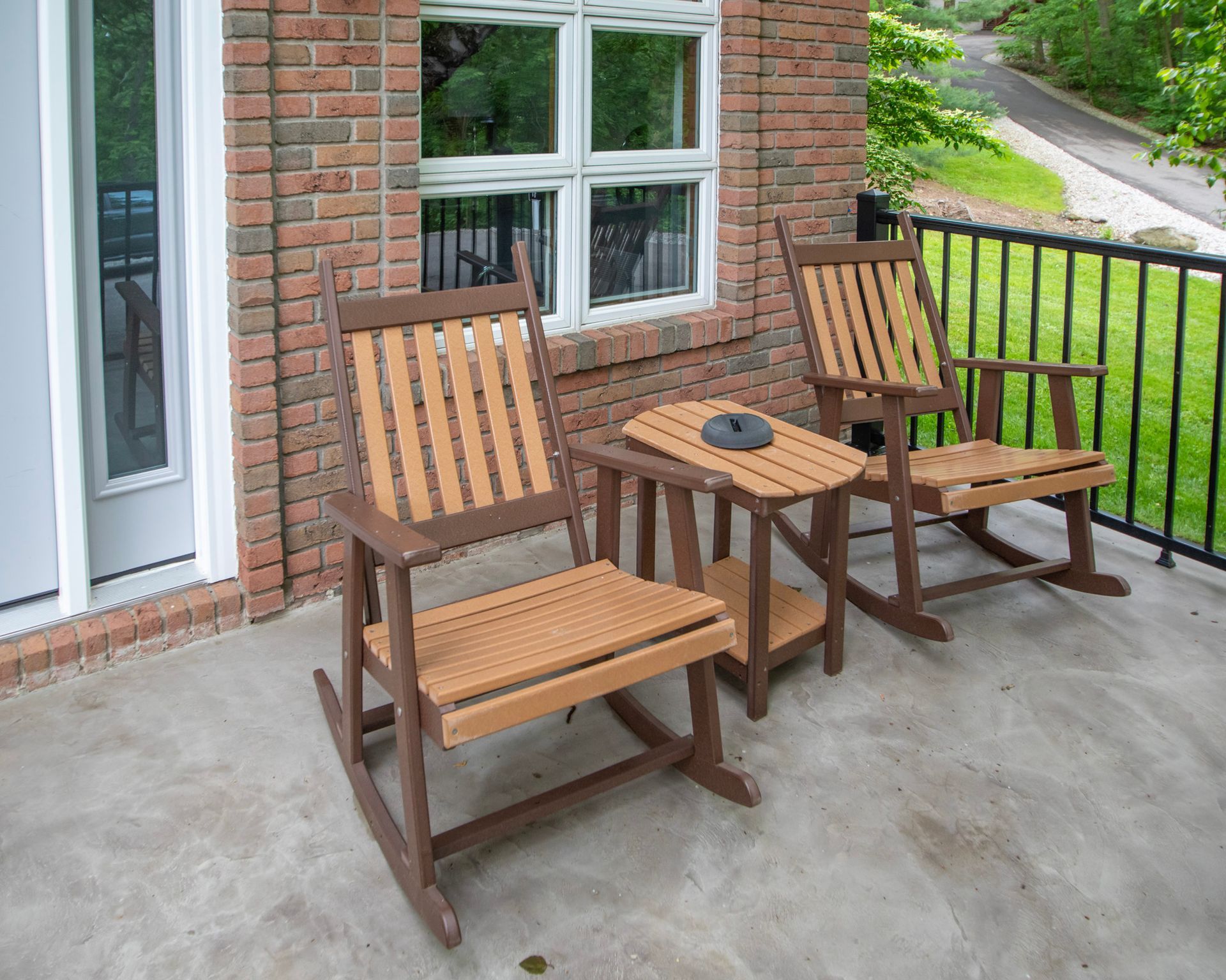 Two wooden rocking chairs and side table on a porch, near a brick wall and window.