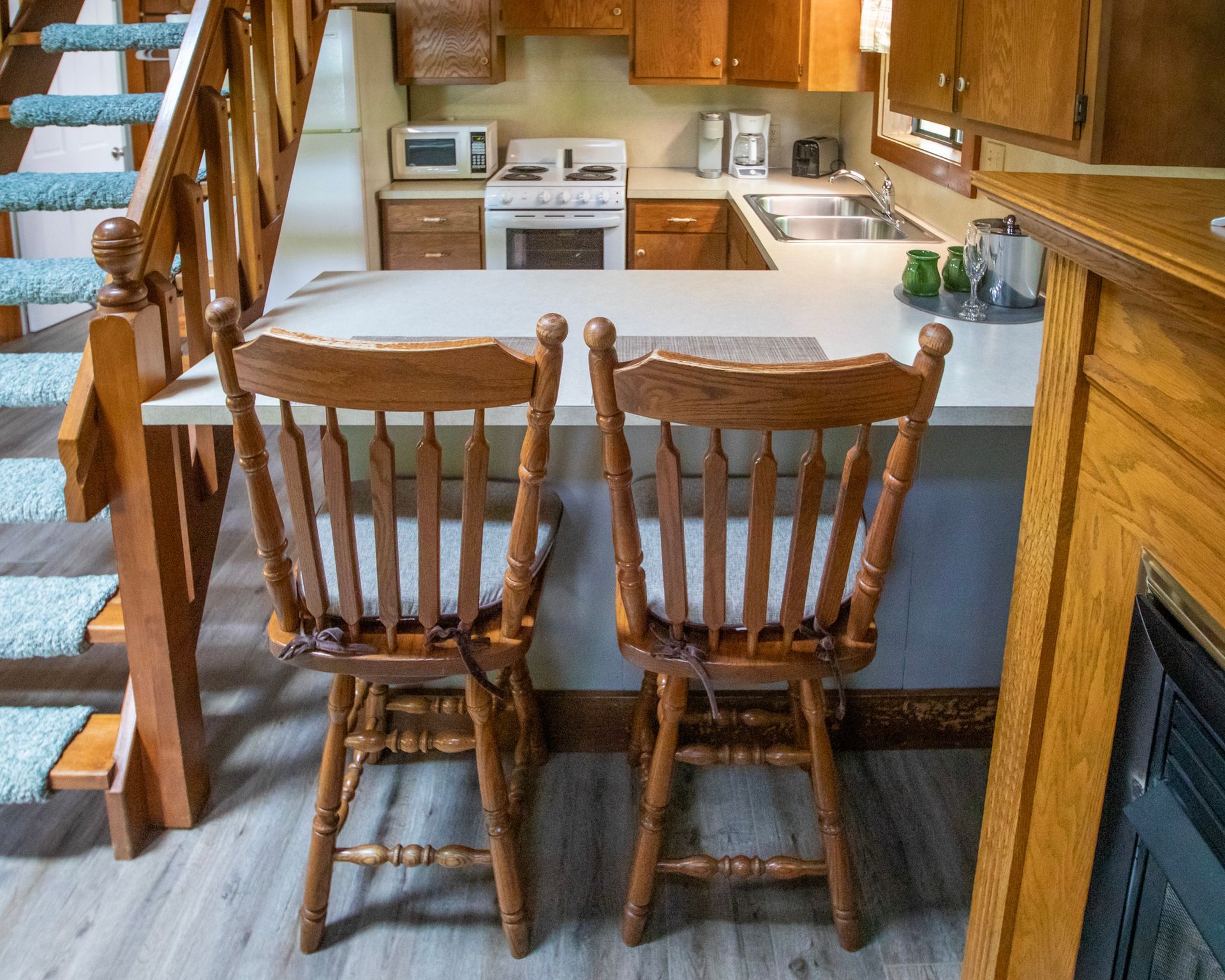 Kitchen with two wooden bar stools at a counter, stairs to the left, and appliances in the background.
