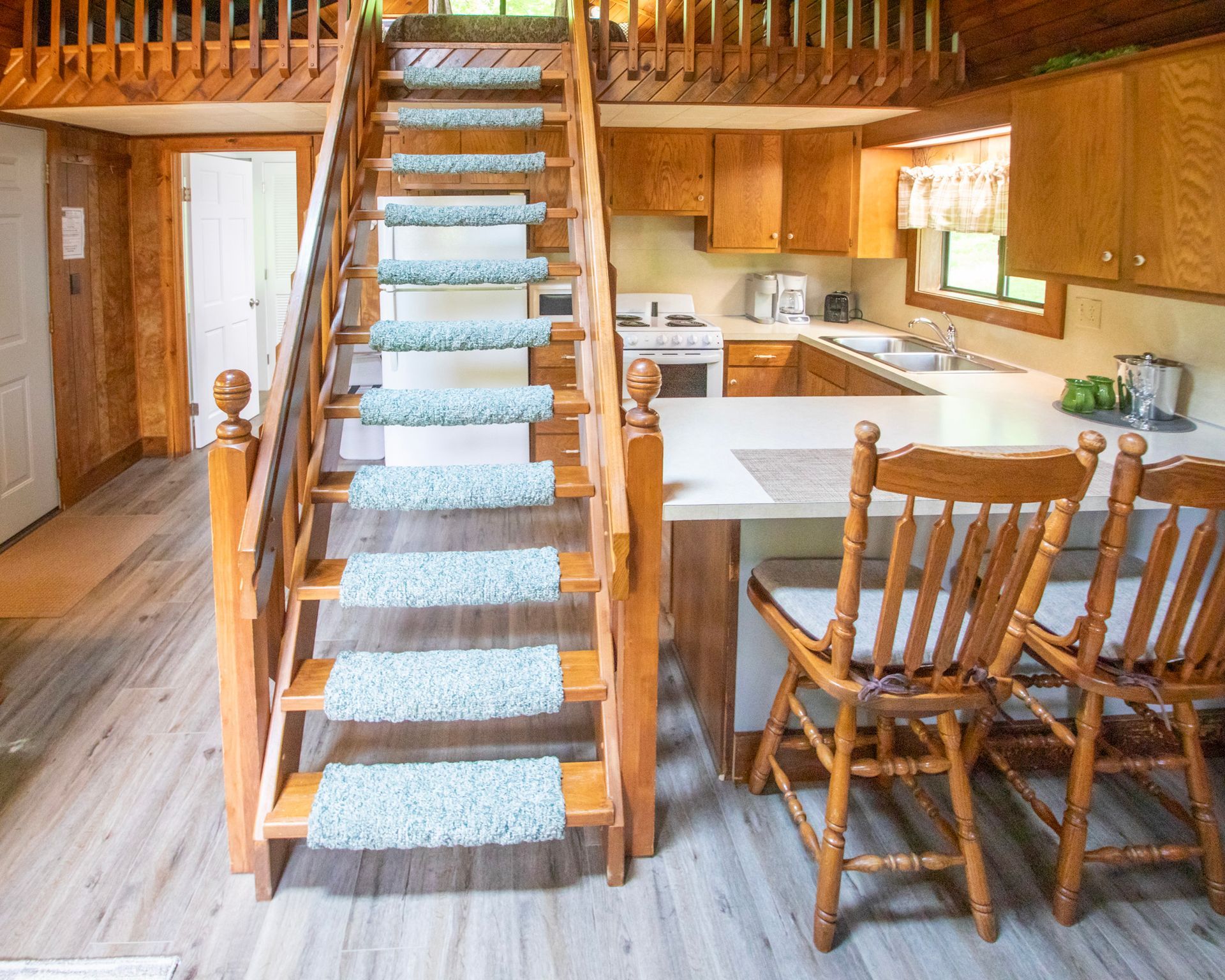 Wooden stairs with blue carpet runners lead to a loft. Kitchen with wood cabinets and dining area are visible.