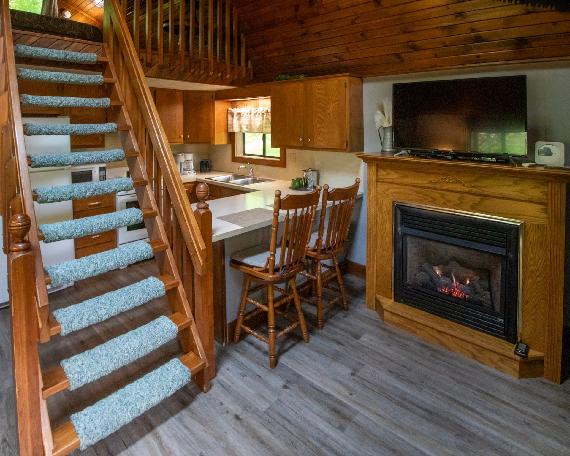 Cozy cabin interior with a staircase, kitchen, dining table with chairs, fireplace, and television.
