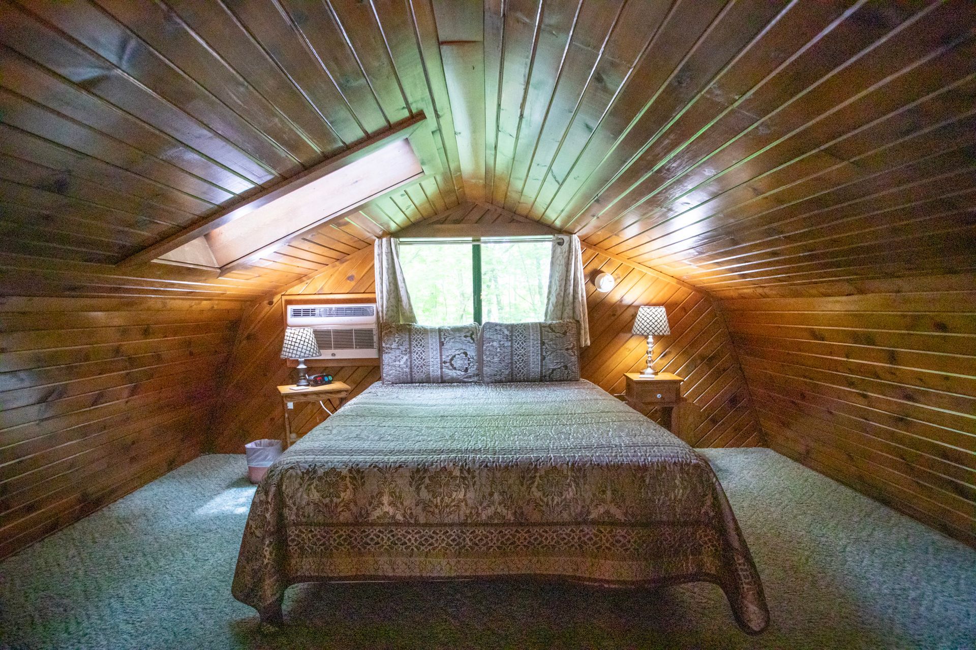Bedroom in a wooden cabin attic with bed, window, and small air conditioner.