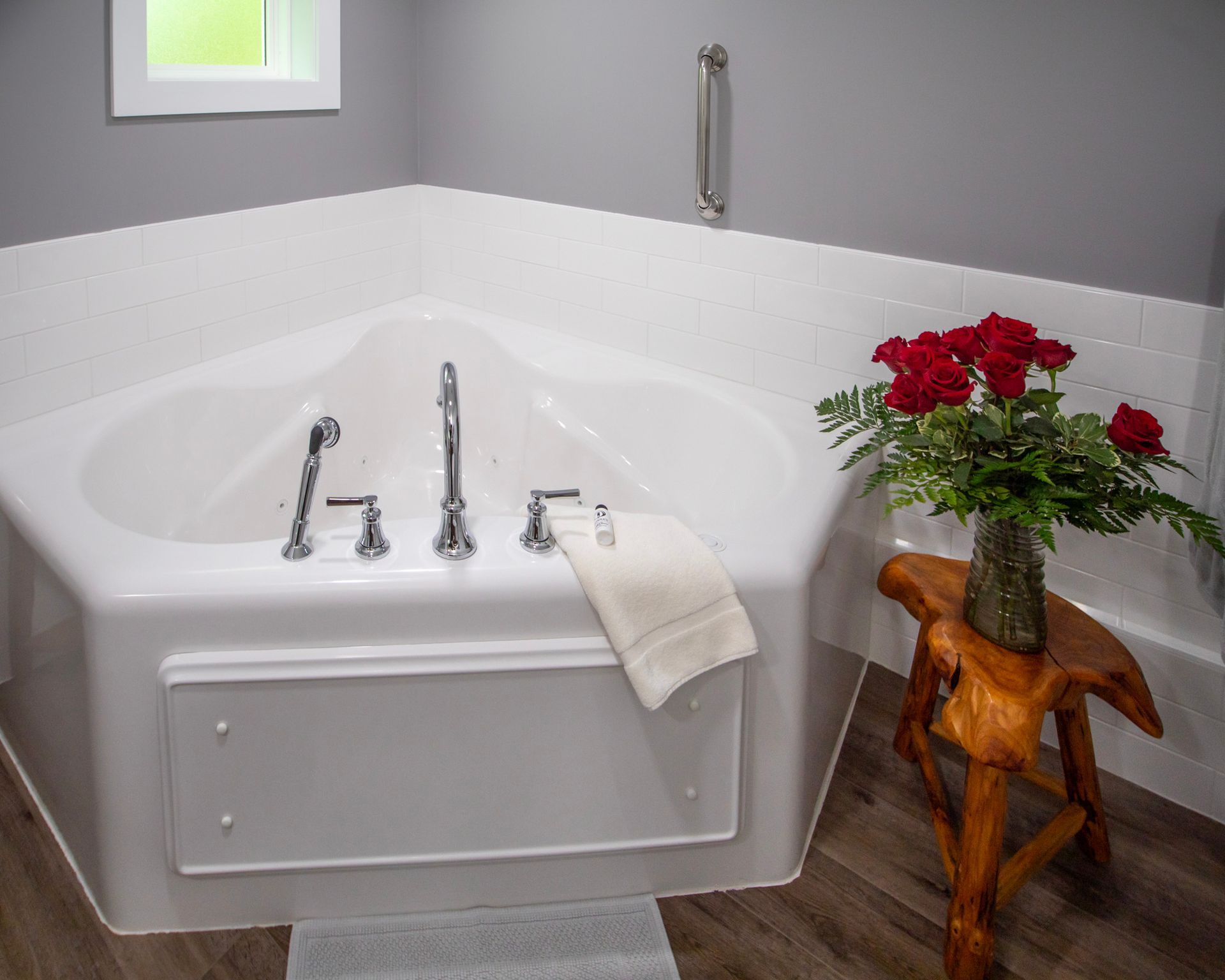 Corner jacuzzi tub in a bathroom, with white tiles, a wooden stool, and a bouquet of red roses.