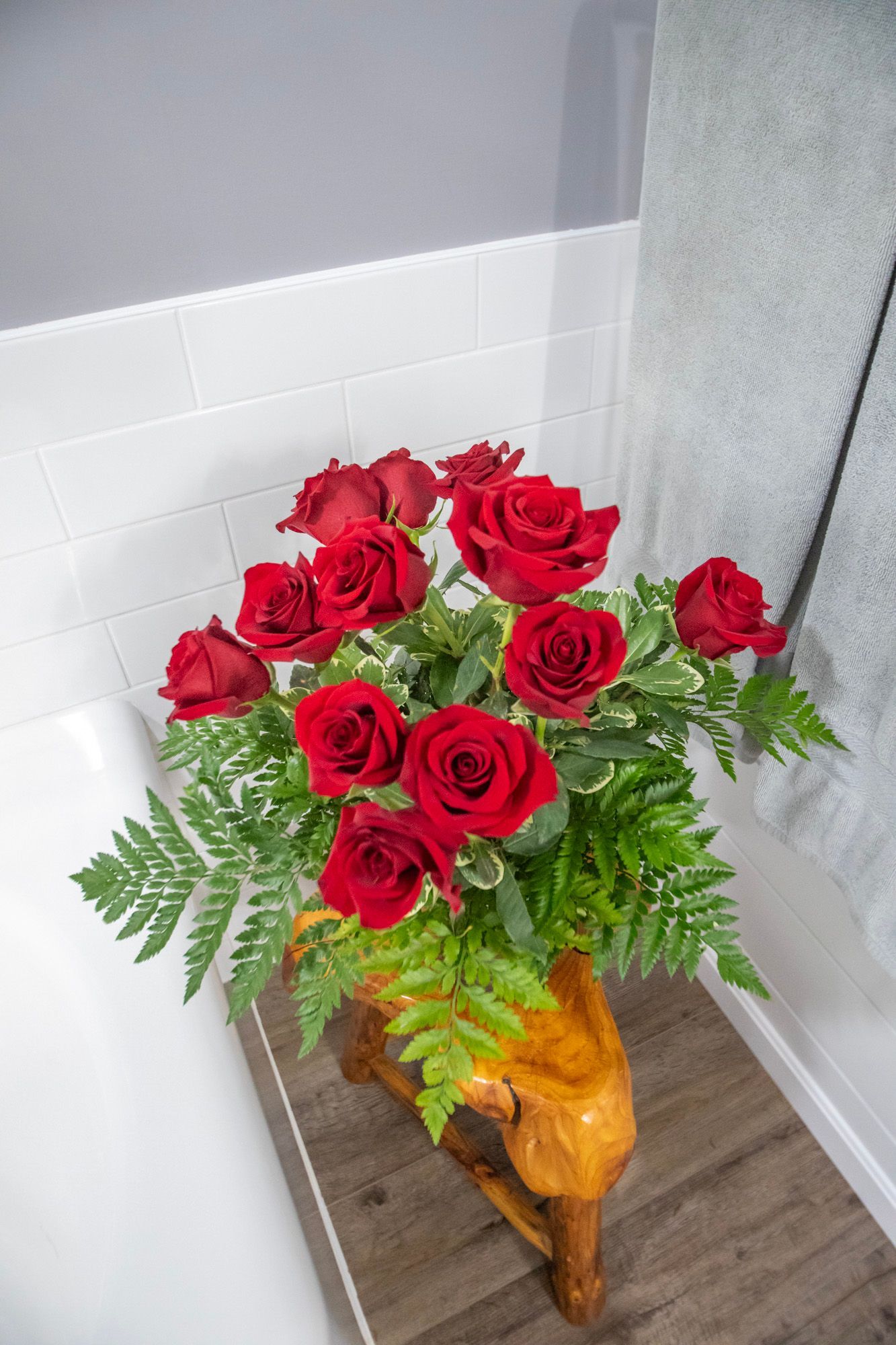 Red roses in a wood-carved vase. Fresh flowers on a wooden shelf, white-tiled wall in background.