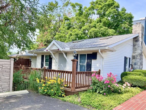 White cottage with a wooden deck, flowers, and a stone chimney.