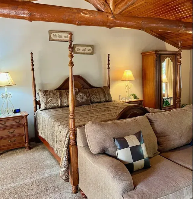 Bedroom with a four-poster bed, nightstands with lamps, a sofa, and an armoire. Warm, cozy setting.
