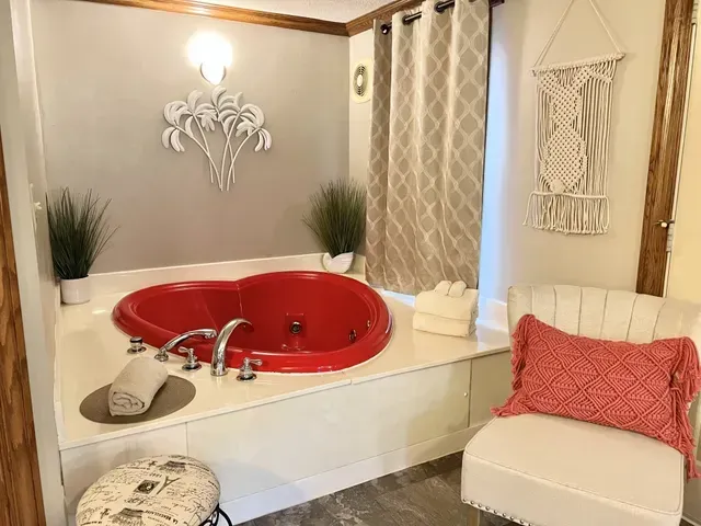 Red heart-shaped jacuzzi tub in a small bathroom, with plants, a chair, and macrame decor.