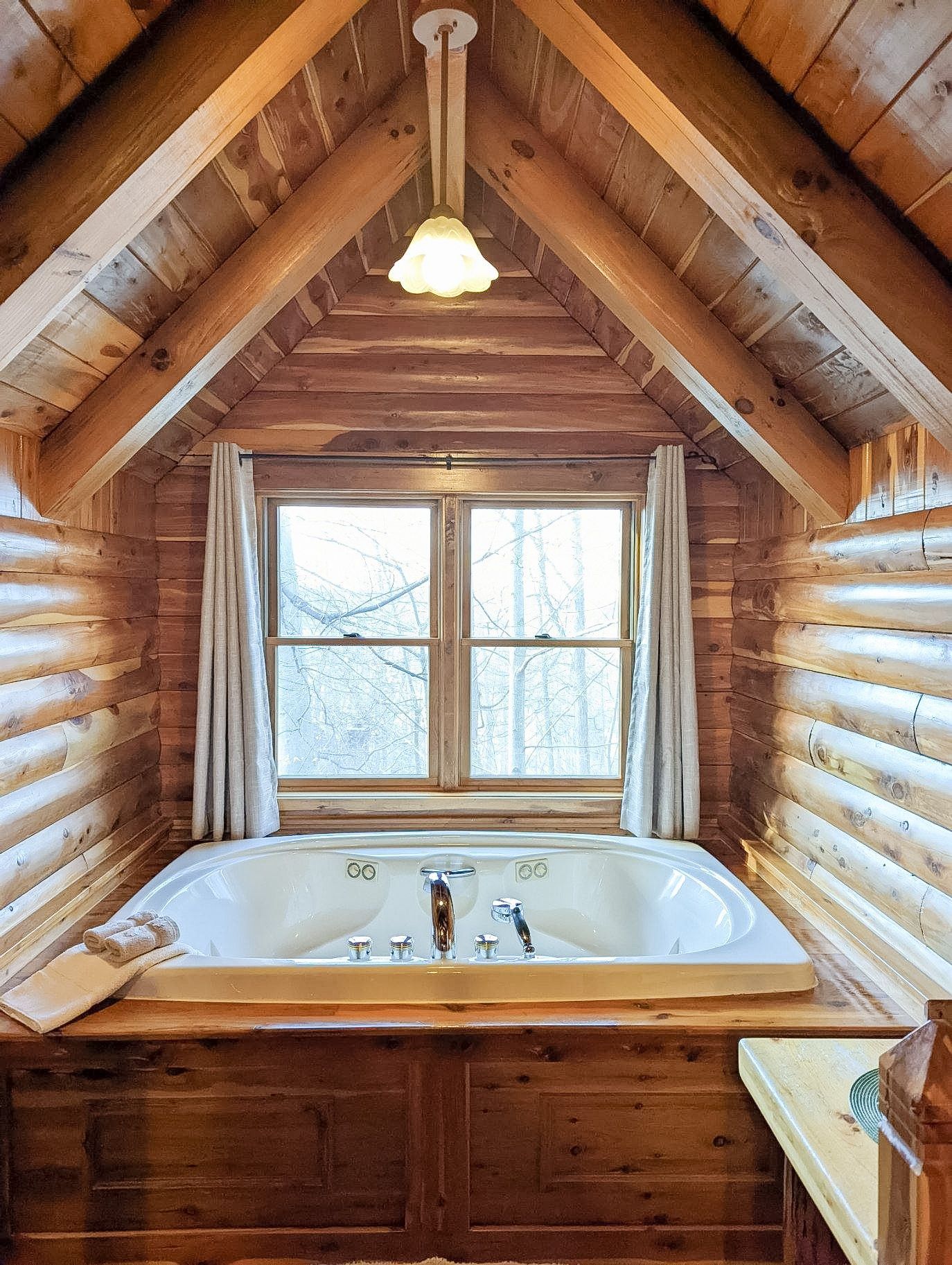 Wooden-walled bathroom with a window and a built-in bathtub. Light shines through, with a hanging light fixture above.