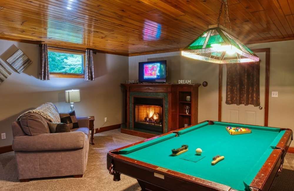 Pool table and fireplace in a basement recreation room. Green felt, wooden paneling, lit fireplace, TV, and sofa.