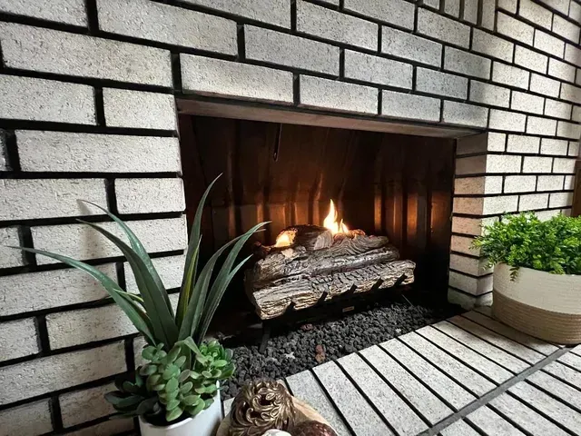 Fireplace with white brick, burning logs, plants in pots.