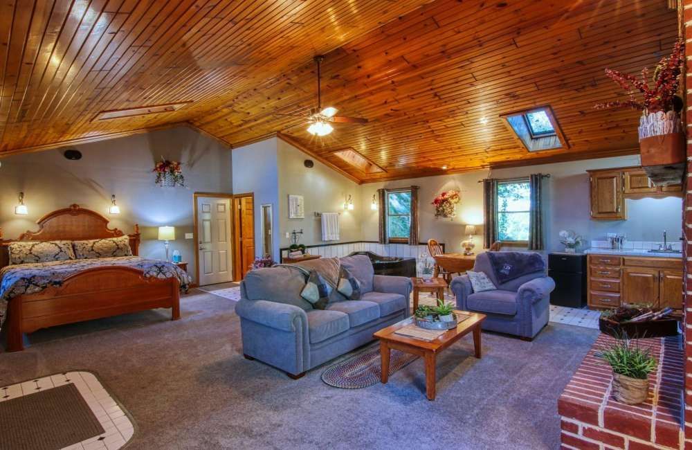Cozy studio apartment with vaulted wood ceiling, bed, sofa, fireplace, kitchenette, windows.