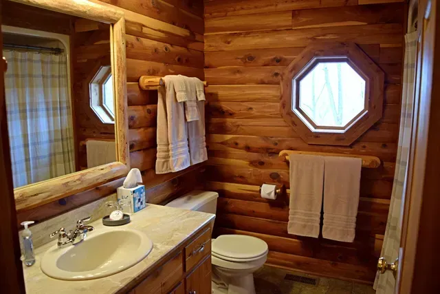 Log cabin bathroom with wooden walls, sink, toilet, and octagonal window. Towels hang on a wooden rack.