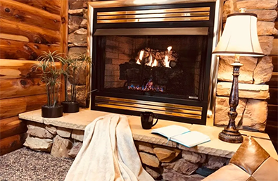 Cozy fireplace scene: fire burning in stone hearth, blanket, open book, lamp, log walls, plants.