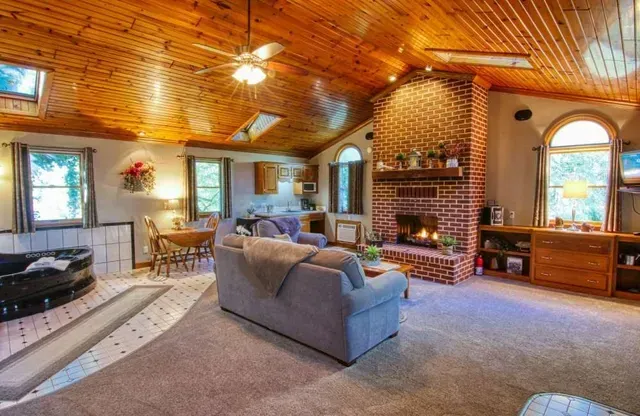 Cozy living room with fireplace, wood ceiling, and windows. Includes seating, dining area, and a jacuzzi tub.