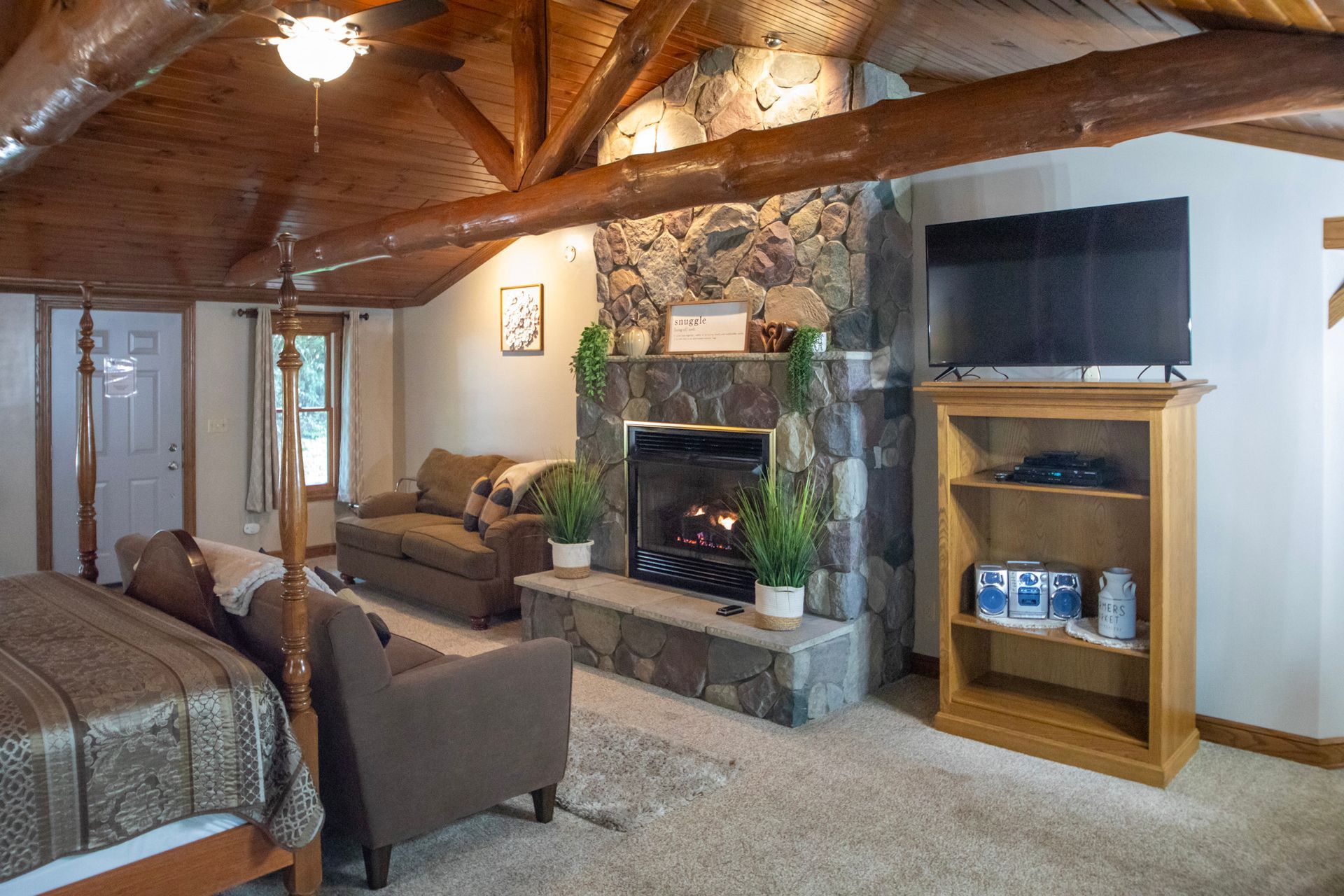 Living room with fireplace, TV, and bookshelves. Features wood beams, a sofa, and plants.