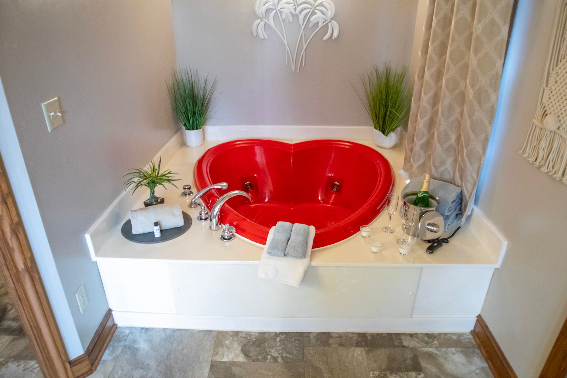 Heart-shaped red jacuzzi tub on white platform, plants, champagne, and curtains.