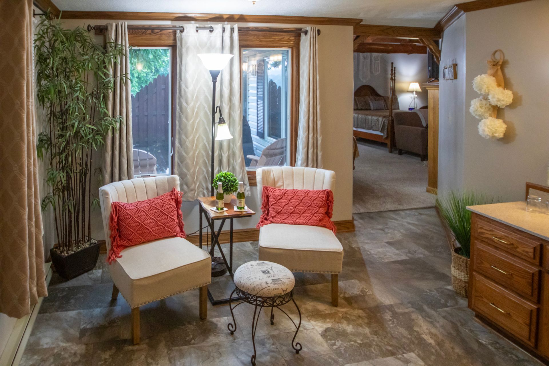 Cozy sitting area with two armchairs, a small table, a stool, and a floor lamp. The room has large windows with outdoor view.