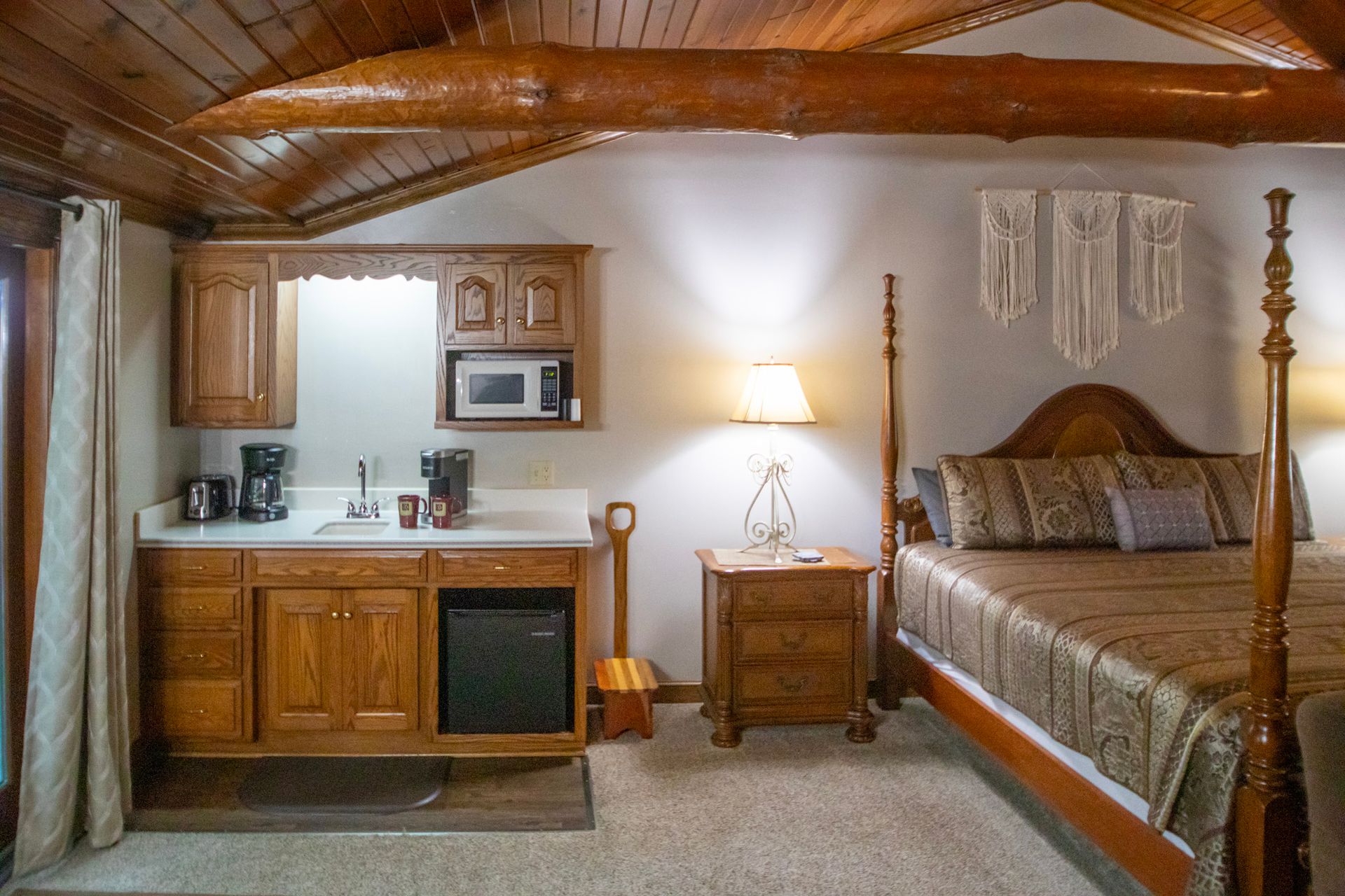 Bedroom with a kitchenette, bed, and nightstand. Wooden accents, beige carpet, and decorative wall hangings.