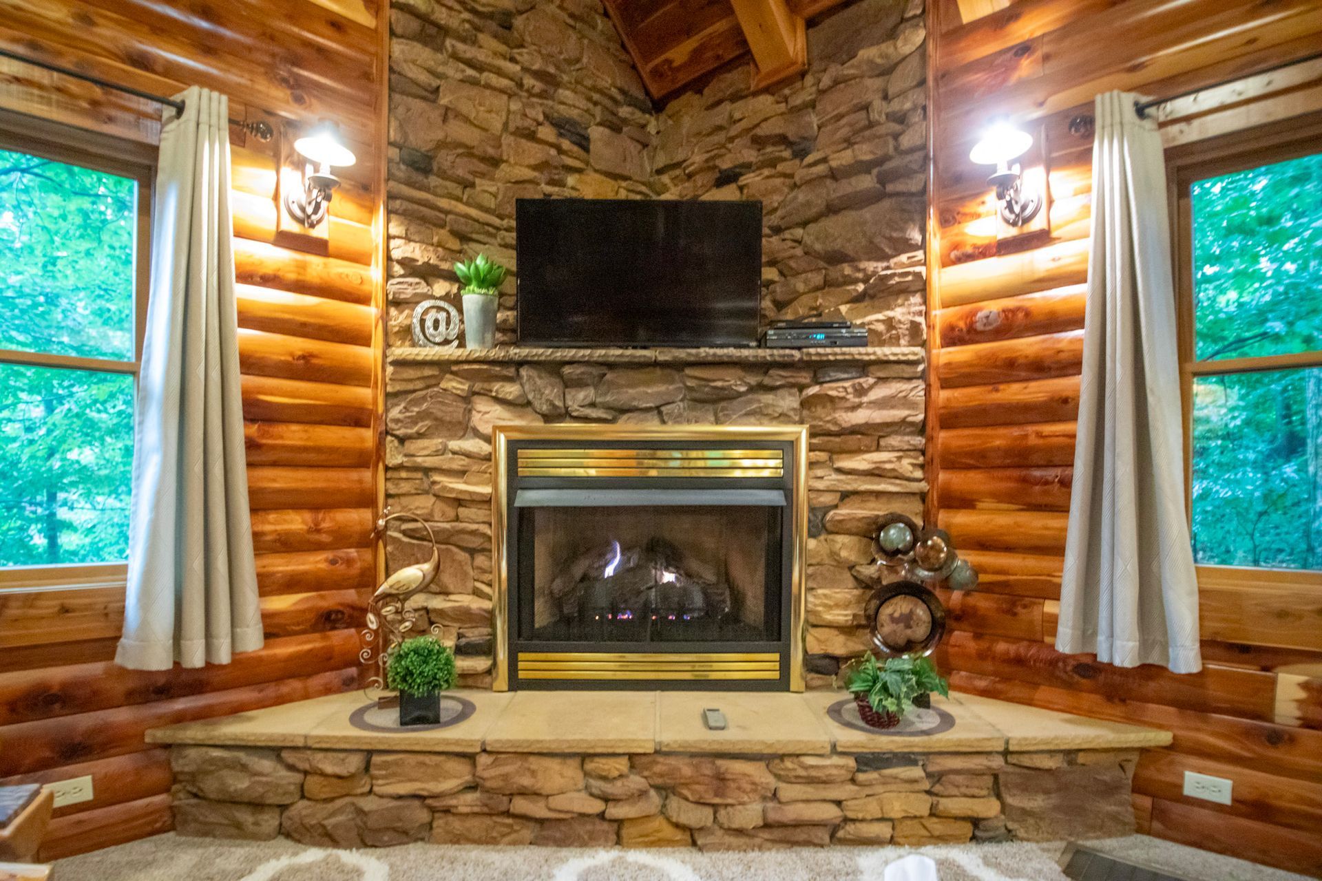Cozy cabin interior with stone fireplace, TV, log walls, and windows with curtains.