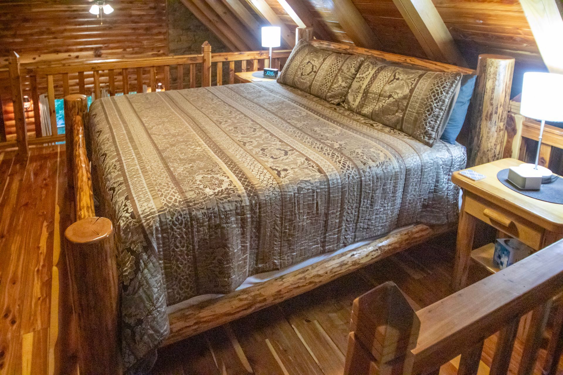 Bedroom with a large bed in a wooden cabin. Bed has a patterned cover. Small table and lamps on the side.