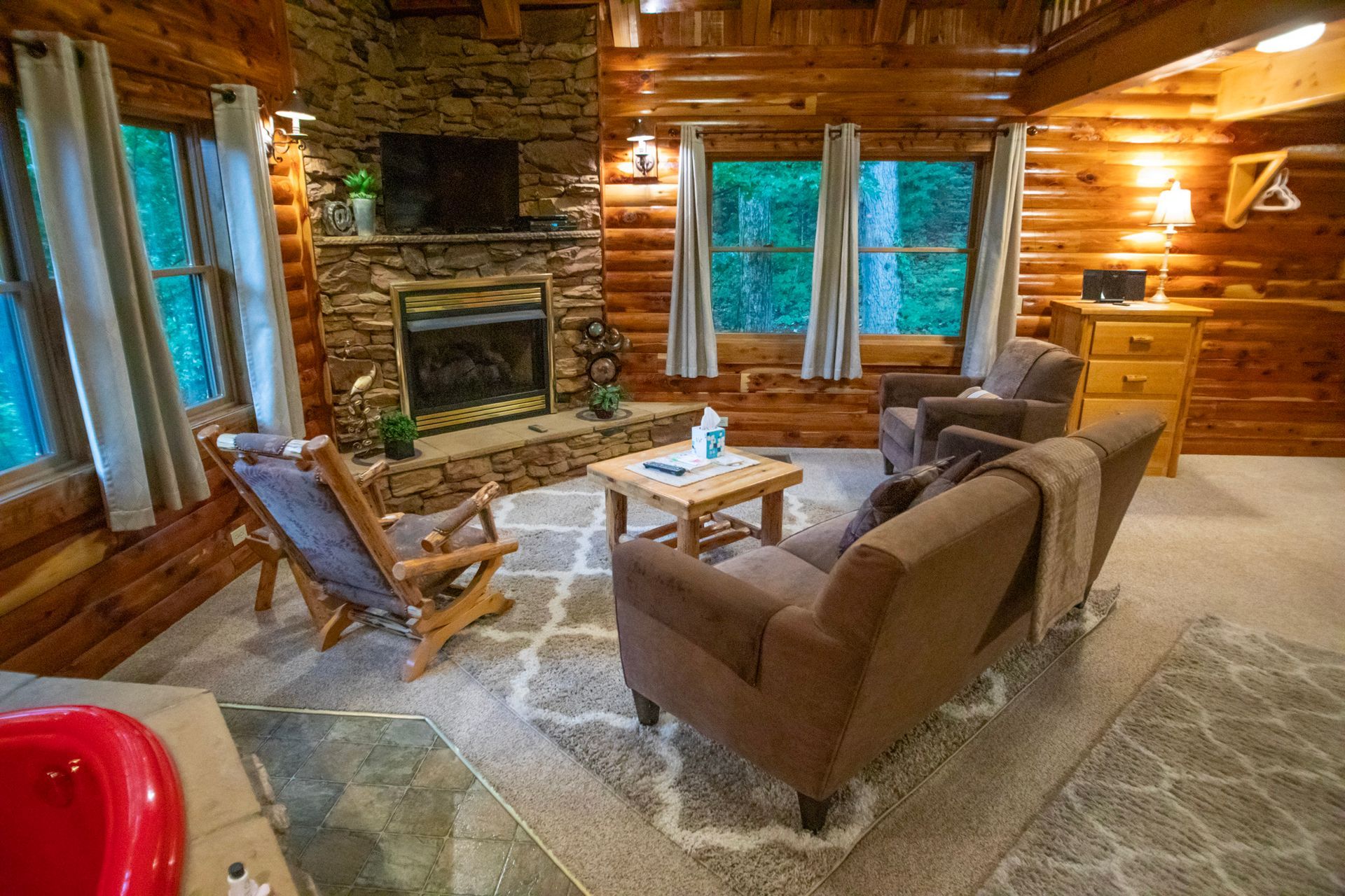 Cozy cabin living room with stone fireplace, log walls, seating, and windows overlooking greenery.