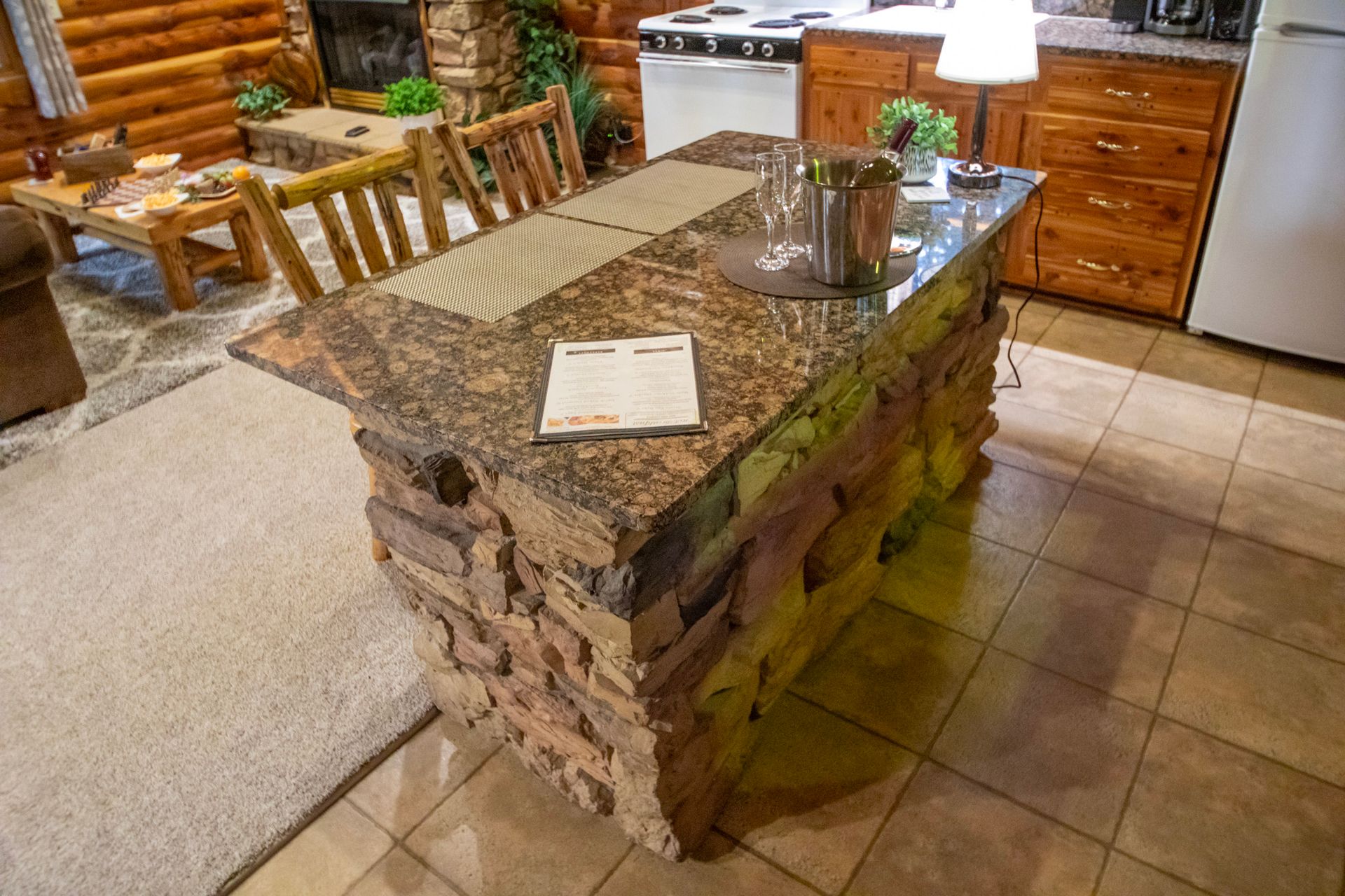 Kitchen island with stone facade and granite countertop, next to dining area in a log cabin.