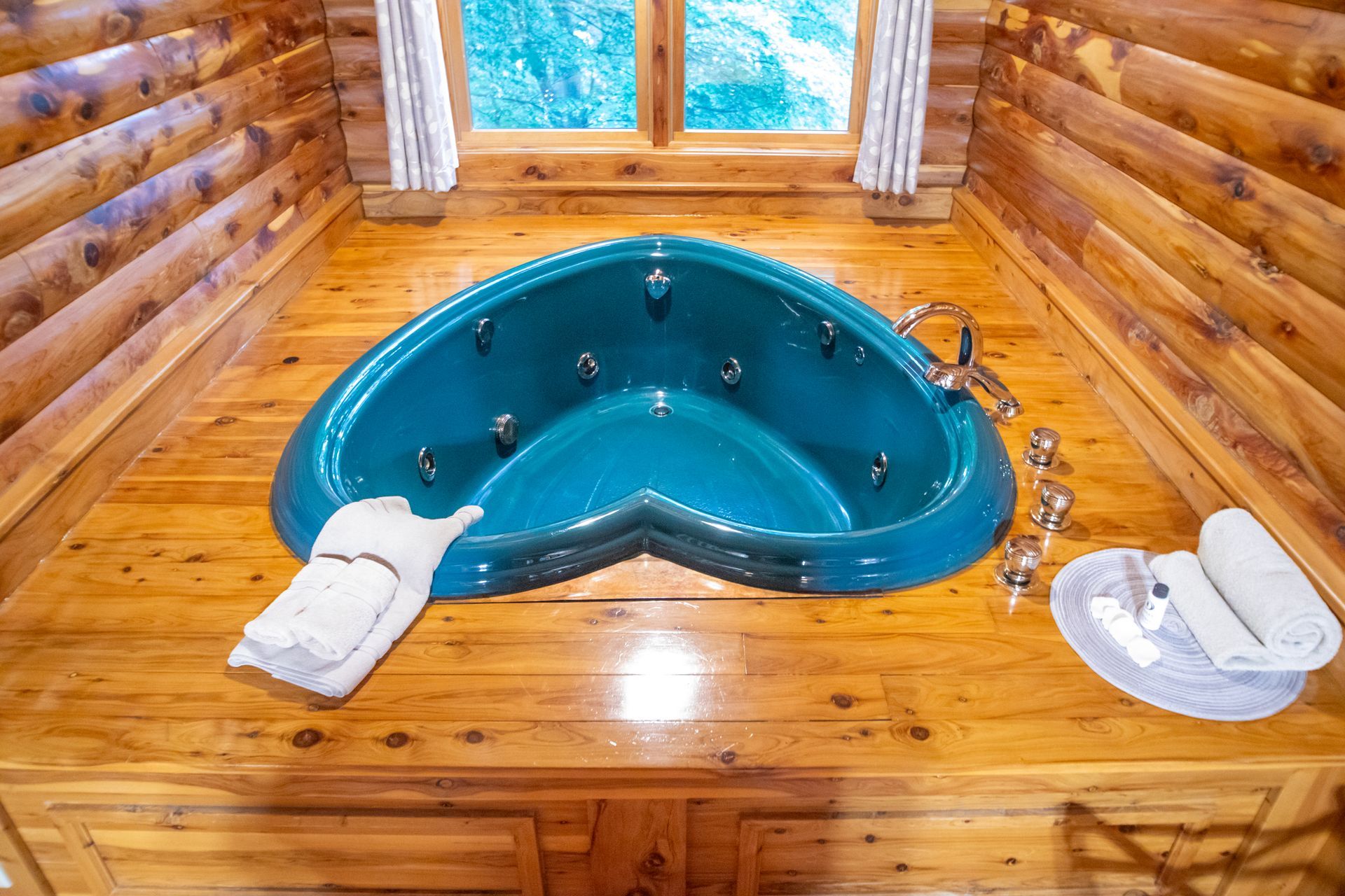 Heart-shaped teal jacuzzi tub in wooden cabin setting, window in background, towels nearby.
