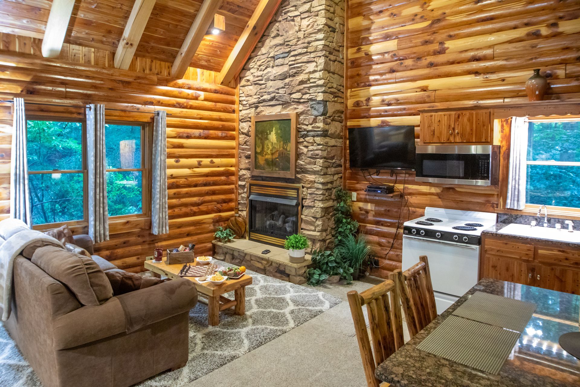 Cozy log cabin interior with fireplace, sofa, kitchen, and windows overlooking greenery.