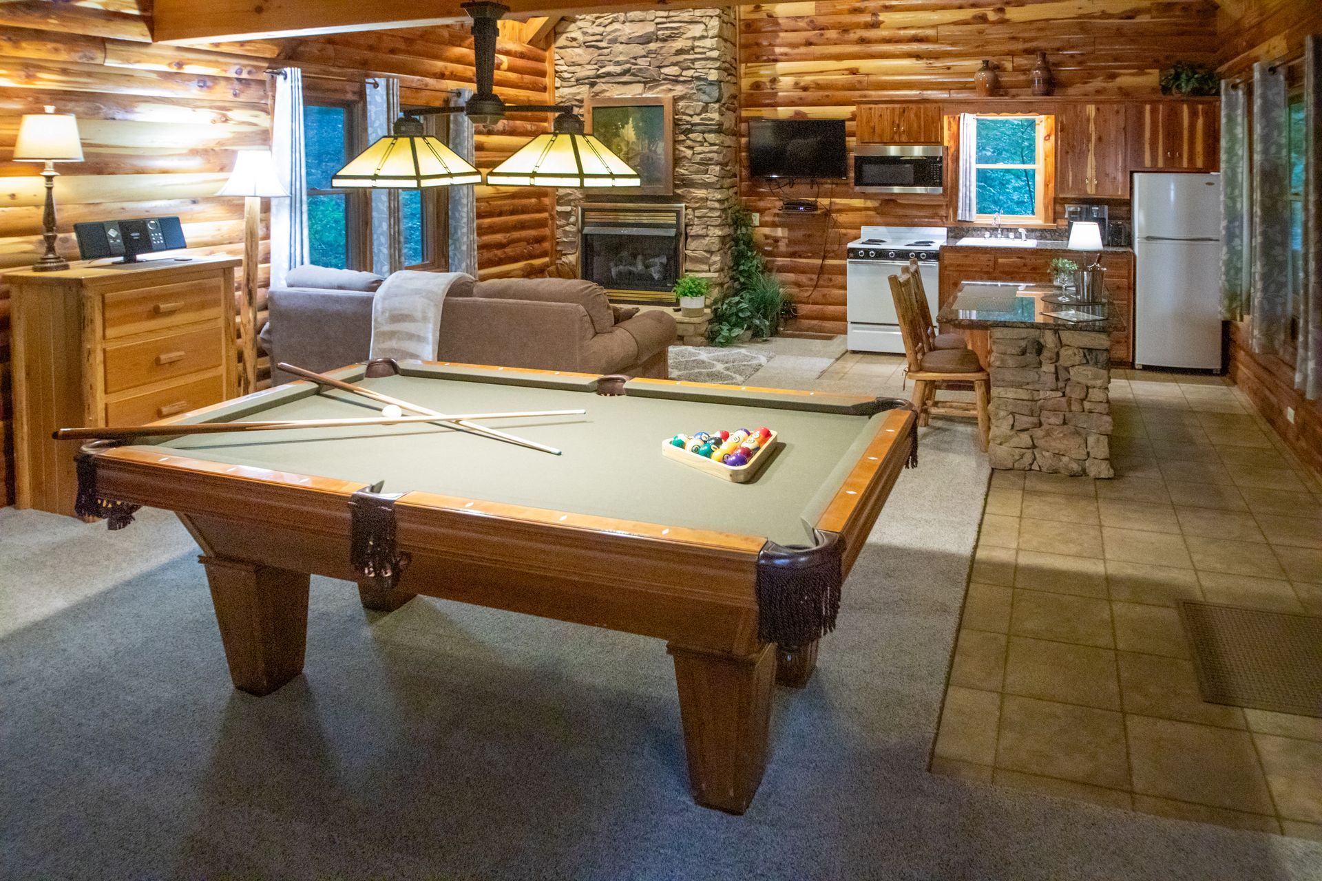 Pool table in a log cabin with fireplace, kitchen, and seating area.