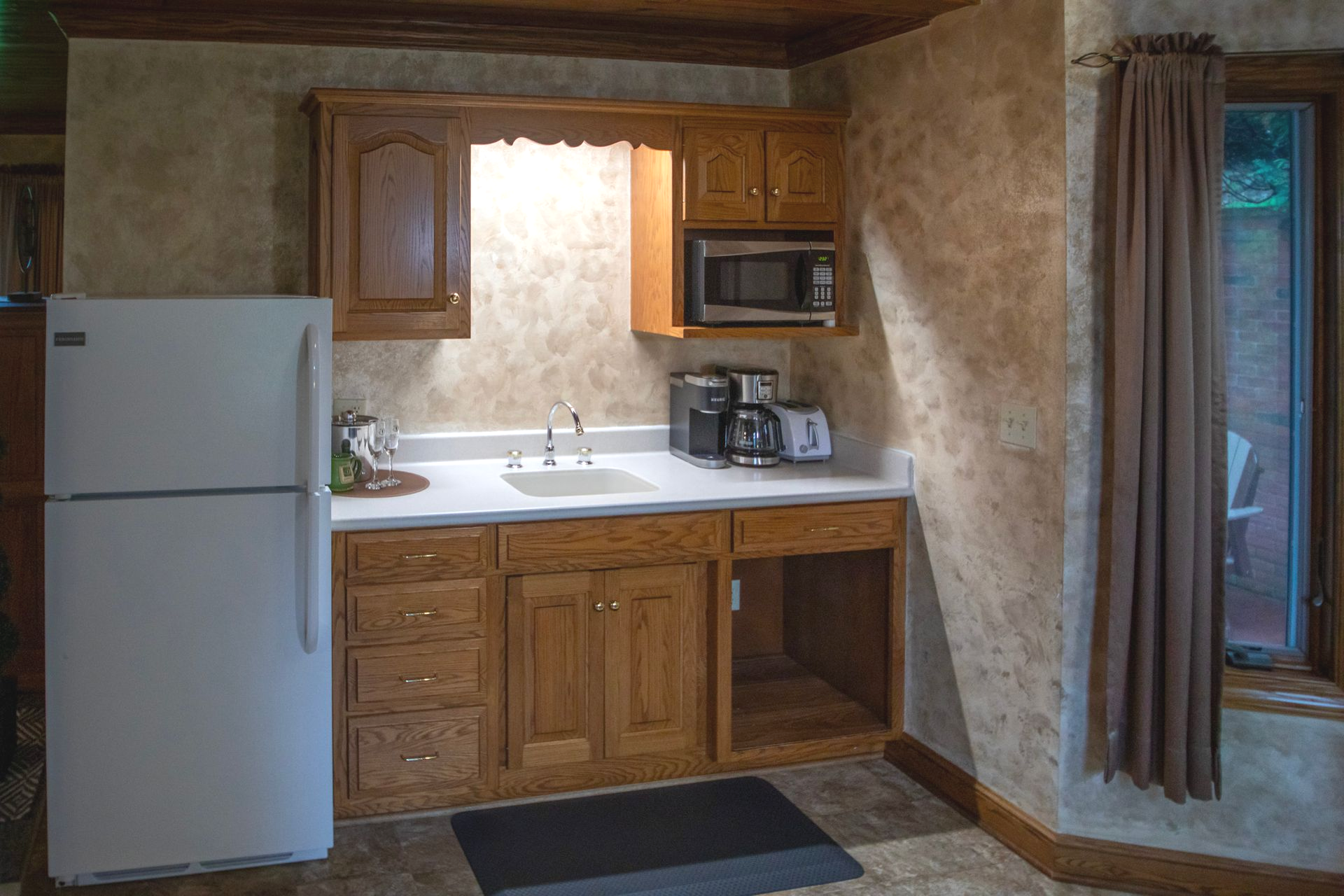 Small kitchen with refrigerator, cabinets, sink, microwave, and toaster. Brown walls and a window with curtains.