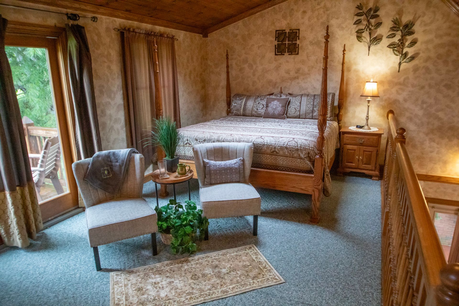Bedroom with wooden four-poster bed, two chairs, and a door to a balcony. Warm tones, carpeted floor.