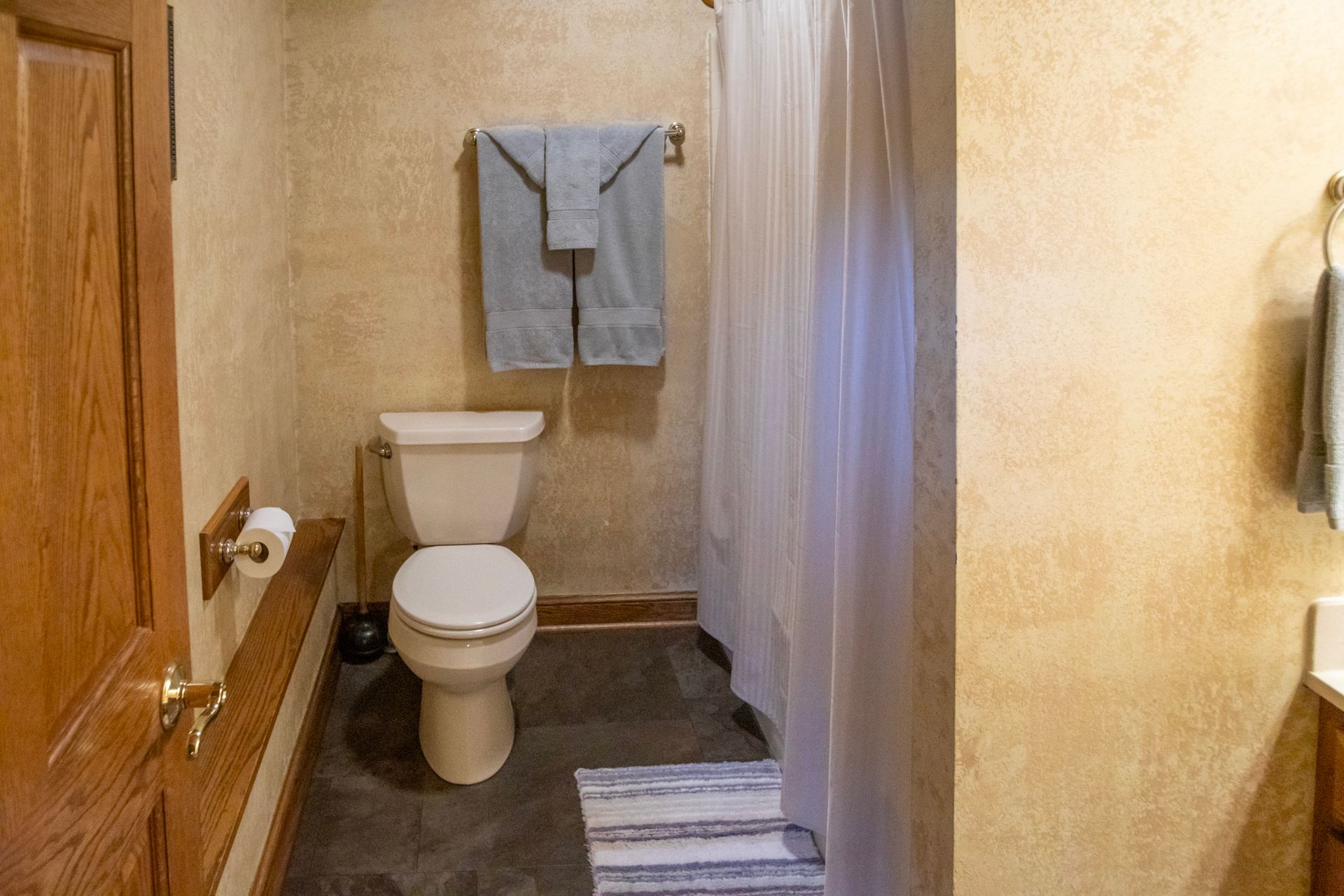 Bathroom with tan textured walls, toilet, shower curtain, and a towel rack with folded towels.