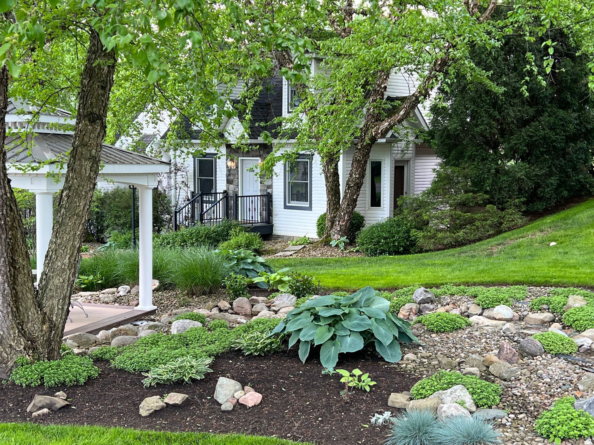 White house with lush landscaping, including trees, shrubs, and a birdhouse; a grassy hill in the background.