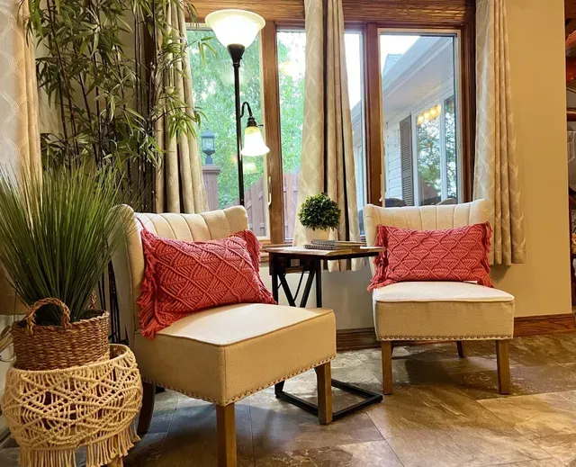 Two beige armchairs with red pillows face a window, next to a lamp and potted plants.