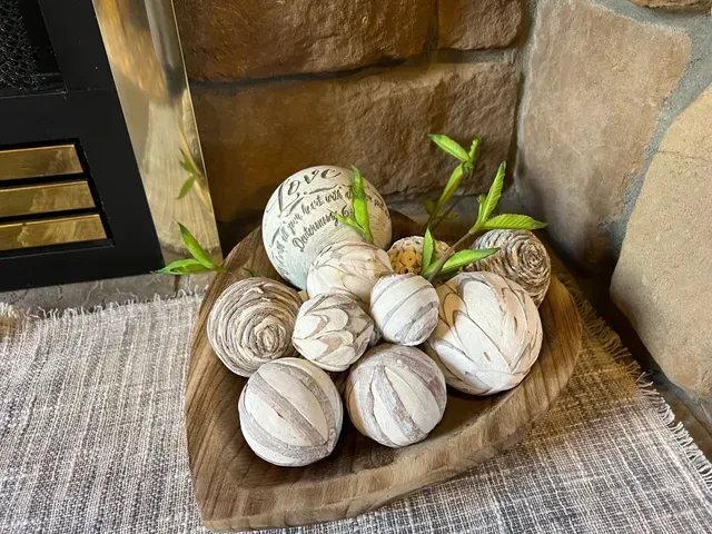 Wooden bowl with decorative spheres, faux greenery, set on a textured rug, near a stone wall.