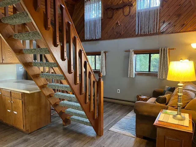 Wooden staircase leads up to a loft area. Brown sofa, windows, and wood paneling in the background.