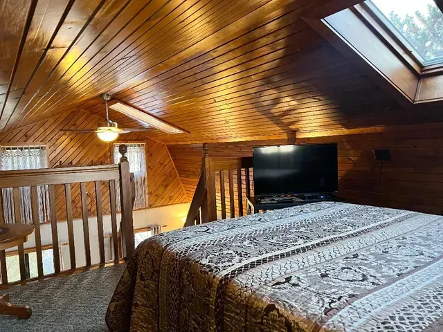 Bedroom loft with wood panel ceiling, bed with decorative spread, television, and skylight.