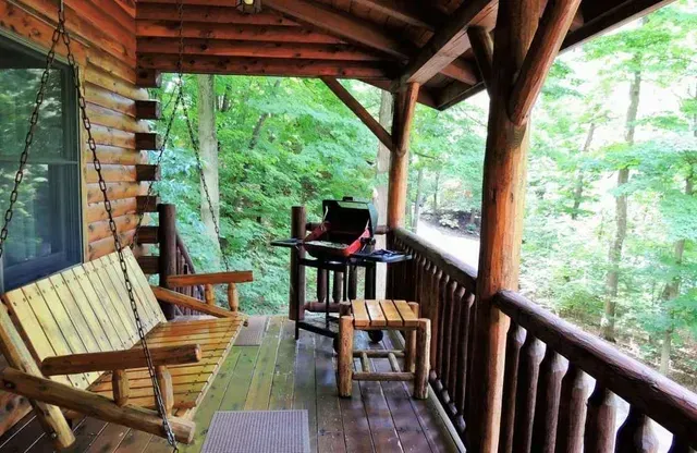 Wooden porch with swing, grill, and small table overlooking a green forest.