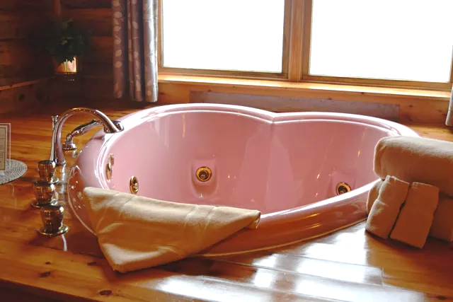 Pink heart-shaped hot tub in a wooden-paneled room, towels nearby, and a window in the background.