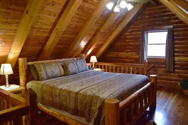 Bedroom in a log cabin with a wooden bed, walls, and ceiling, a window, and two lamps.