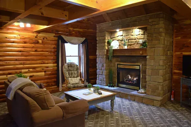 Cozy living room with a stone fireplace, log walls, and a window. A couch and armchair are present.