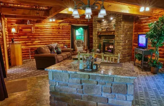 Cozy cabin interior with stone fireplace, log walls, and a granite-topped island; sofa and television are visible.
