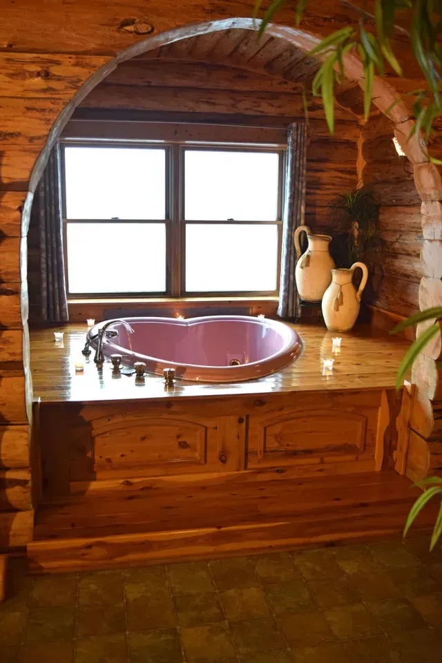 A pink jacuzzi tub in a wooden alcove with a window. Two vases and greenery are on the wall.