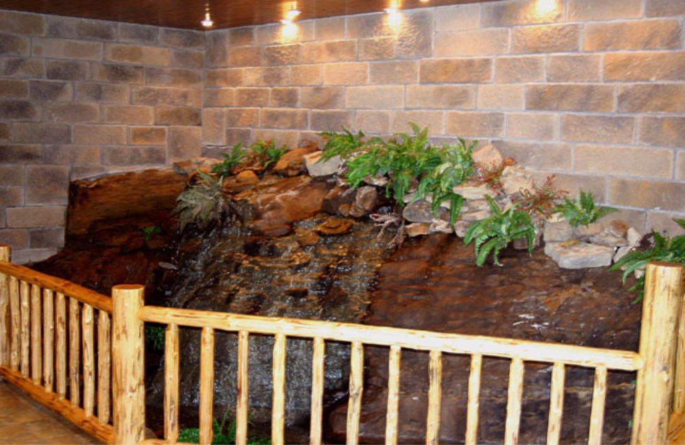Indoor rock waterfall with greenery, brick wall backdrop, and log railing.