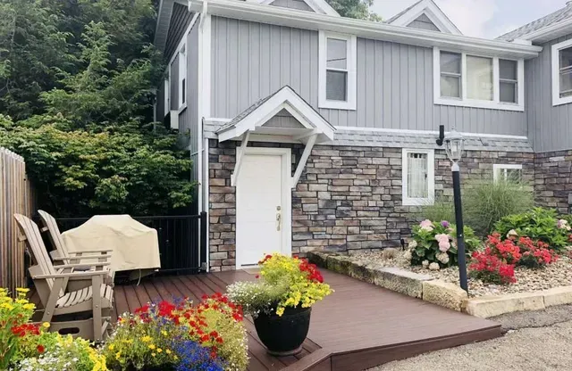 Townhouse exterior with stone accents, gray siding, white door, and flower-filled planters on a wooden deck.