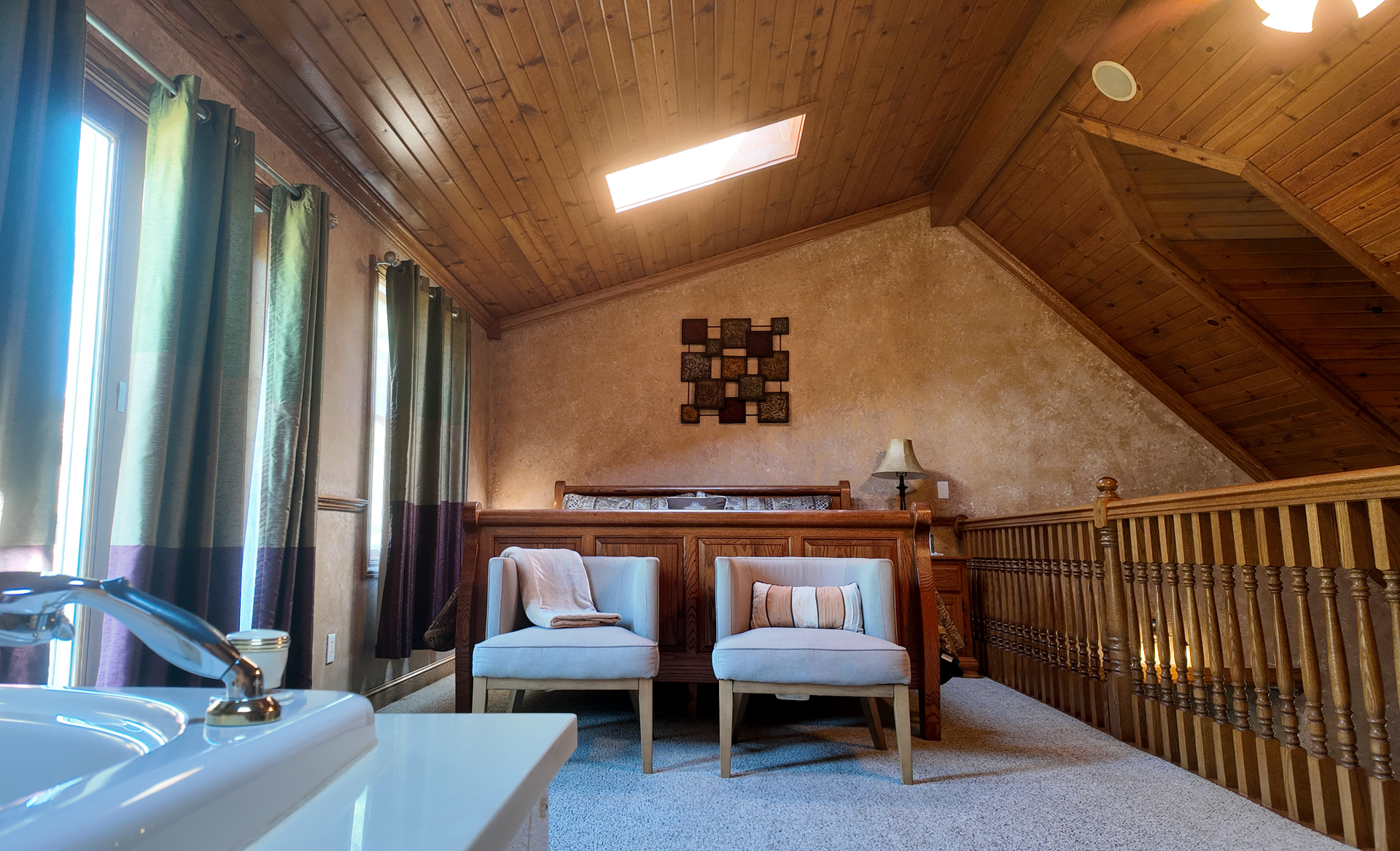 Loft bedroom with wooden accents, two chairs, a jacuzzi tub, and a window with curtains.