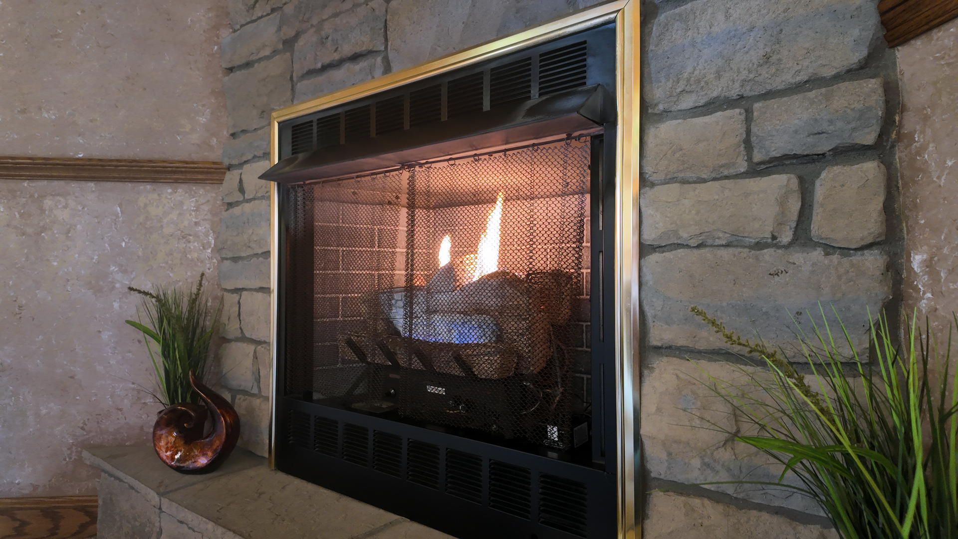 Fireplace with flames behind a mesh screen, surrounded by stone and a gold frame.