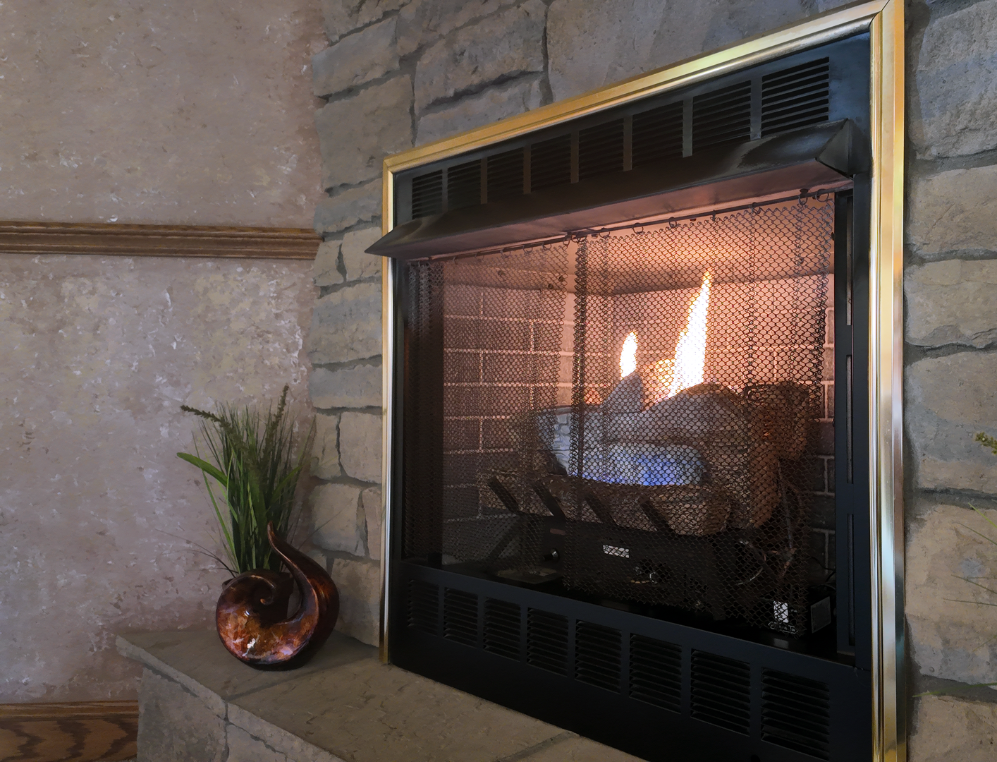 Fireplace with flames behind a mesh screen, stone wall background, and decorative vase.