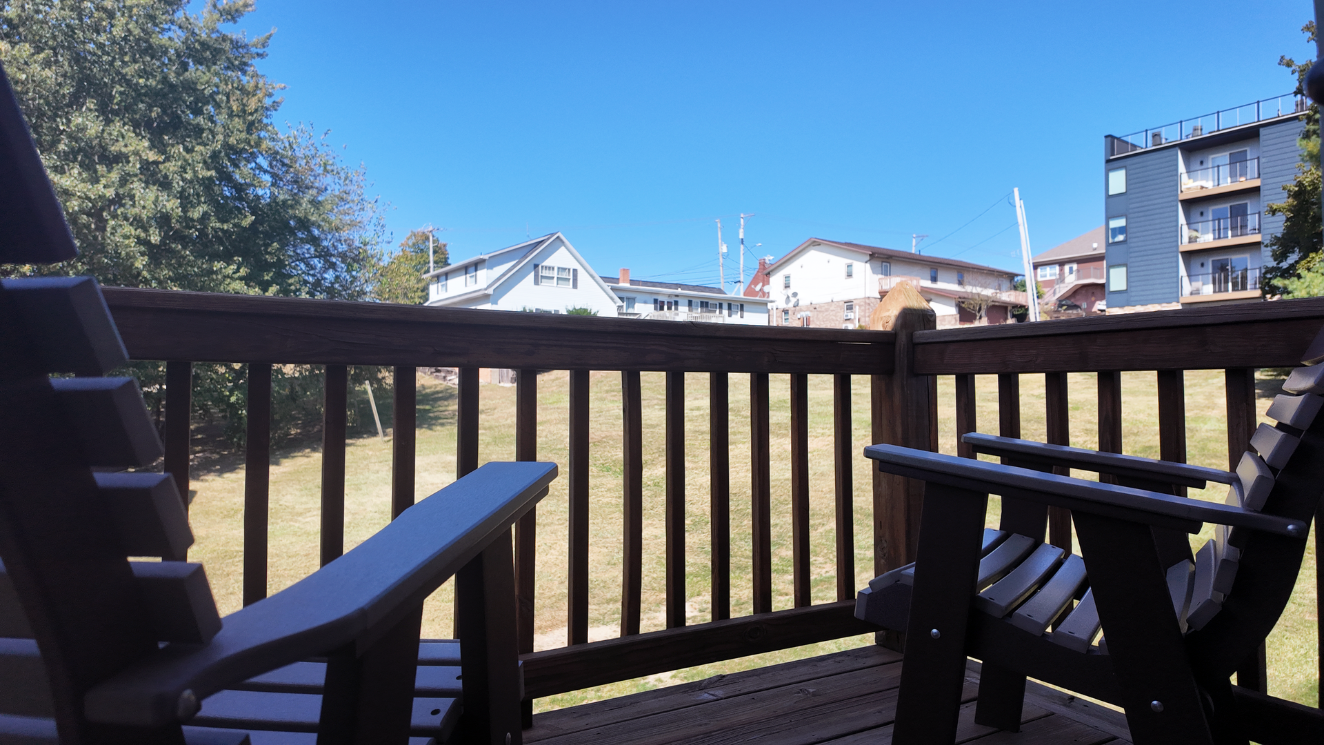 Two wooden Adirondack chairs sit on a deck overlooking a yard and houses under a clear blue sky.